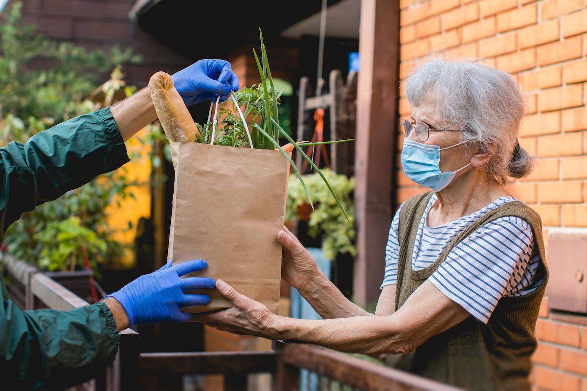 Senior woman wearing a face mask receiving a paper bag of groceries from a gloved delivery person outdoors, highlighting contactless meal delivery service in Ottawa.