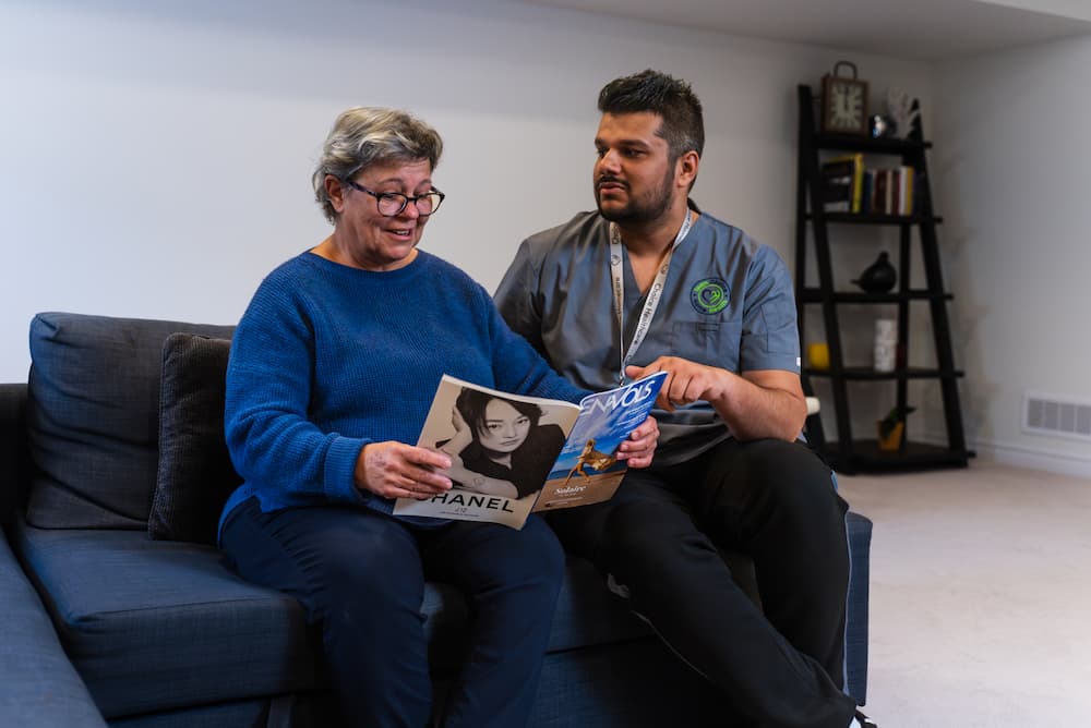 A male nurse having a conversation with an elderly woman who is reading a magazine